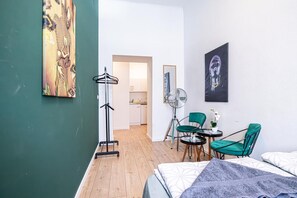 A hotel room with a bed, two chairs, a small table, a fan, and artwork on the walls.