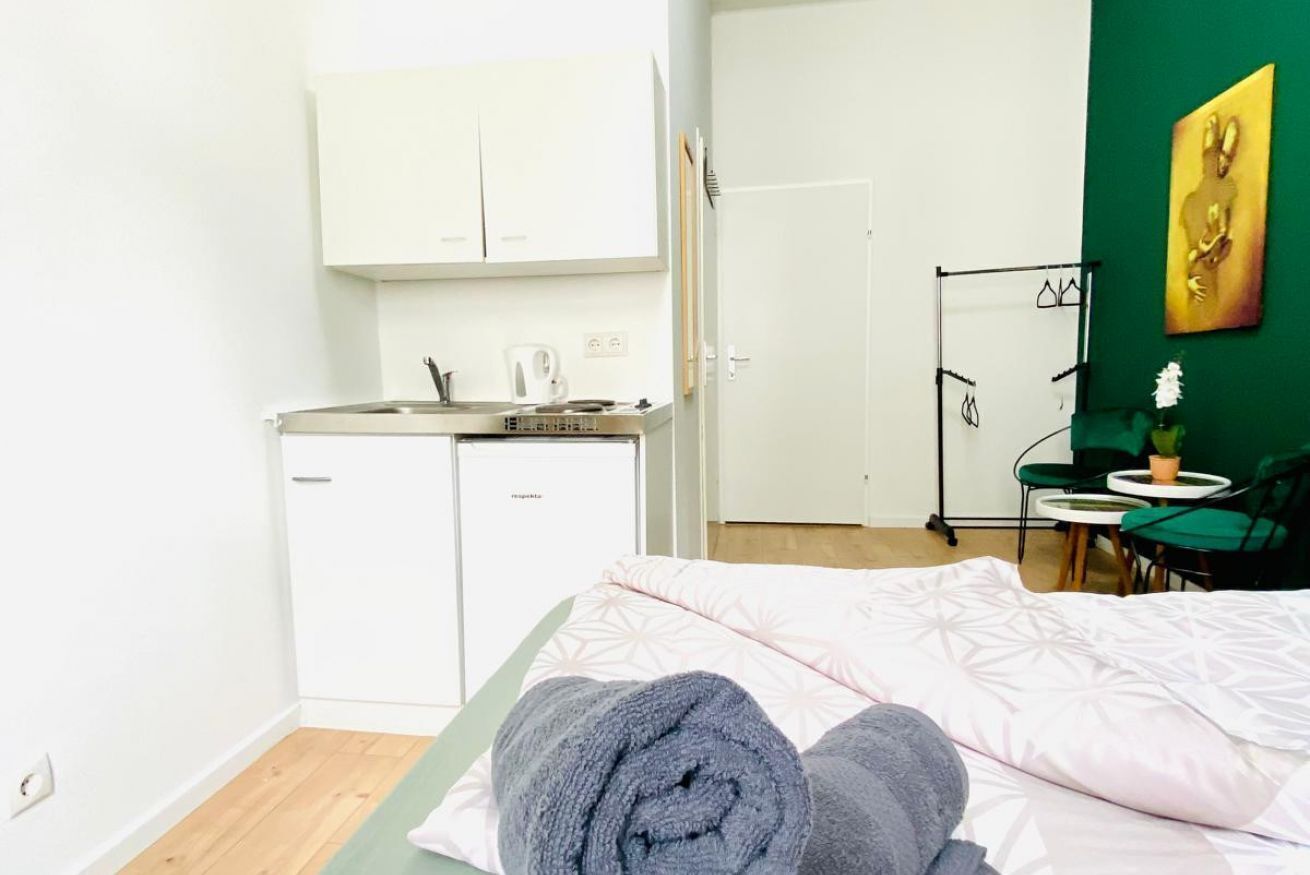 Ein Schlafzimmer mit Bett, gerolltem Handtuch, einer kleinen Küchenzeile und einer grünen Wand mit einem Gemälde.