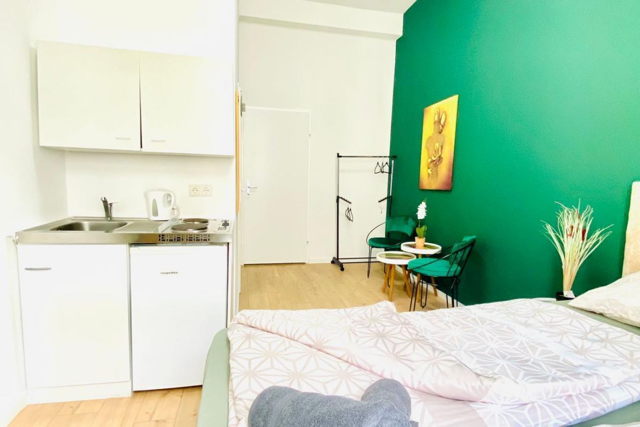 A compact room with a kitchenette, a bed with a patterned bedspread, a green accent wall, and a small dining area.