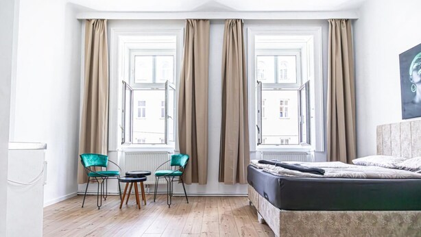 Ein Schlafzimmer mit einem Bett, zwei mit Vorhängen versehenen Fenstern, einem Sessel, einem kleinen Tisch und einem Heizkörper.