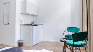 Eine kleine Küchenzeile mit Kühlschrank, Spüle und Mülleimer. An der Wand befindet sich ein Spiegel und ein Sessel mit Kissen.