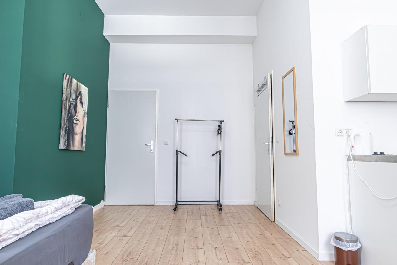 Ein Zimmer mit einem Bett, einer Tür, einem Metallregal, einem Spiegel und einer Mülleimer.
