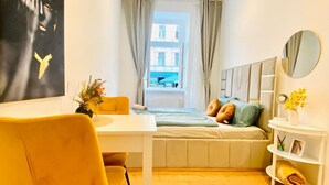 A compact room with a bed, a desk, a chair, a window with curtains, a mirror, and a wall-mounted shelf. - Cozy Room in Ottakring: Spectacular City Views (Vienna)