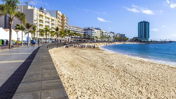 A beachside promenade with a sandy beach, palm trees, and a row of multi-story buildings. - Eslanzarote Wanderlust house (Arrecife)