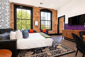A modern hotel room with a large bed, a brick accent wall, a flat-screen TV, and a view of the city.