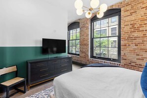 Une chambre à coucher avec un lit, un téléviseur posé sur une commode, un banc et de grandes fenêtres donnant sur un mur de briques.