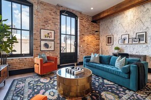 Una sala de estar con pared de ladrillo, ventanas amplias, un sofá azul, un sillón naranja y una mesa de centro redonda.