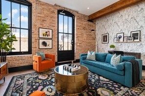 Un salon moderne avec un mur en briques, de grandes fenêtres, un canapé bleu, un fauteuil orange et un tapis à motifs.