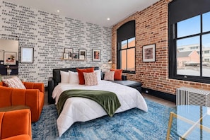 A hotel room with a brick wall, a large bed with a white comforter, orange chairs, and a blue patterned rug. - Spacious Downtown Suite Sleeper Sofa Hotel (New Orleans)