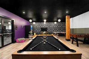 Une table de billard avec ses queues, une table de baby-foot et une table de billard dans une pièce aux murs violets et à un mur orné d’un motif noir.