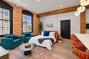 A modern hotel room with a brick wall, large windows, a bed with blue and white bedding, teal and orange chairs, a small round table, and a patterned rug. - Historic Modern Suite Downtown Hotel (New Orleans)