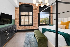 A bedroom with a large bed, a TV, a dresser, and two windows with a view.