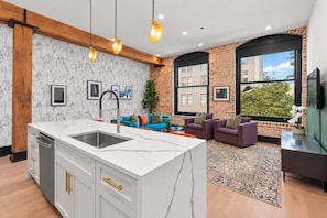 A modern kitchen with a marble island, a built-in oven, and a sink. There is a seating area with a blue sofa and purple armchairs. The room has exposed brick walls and large windows.