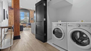 A modern laundry room with two front-loading washing machines, a wooden pillar, and a door leading to a living area with a brick wall and a large window. - Suite Best Location Walk to Everything Downtown Hotel (New Orleans)