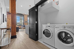 A modern laundry room with two front-loading washing machines, a wooden pillar, and a door leading to a living area with a brick wall and a large window. - Suite Best Location Walk to Everything Downtown Hotel (New Orleans)