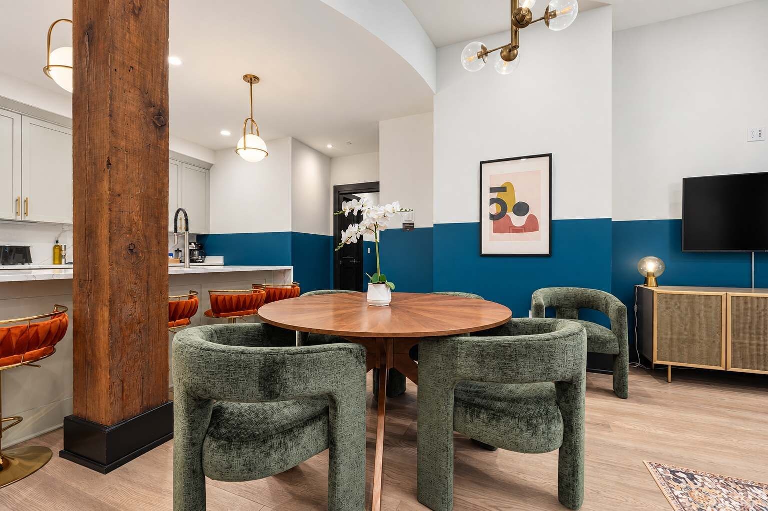 Un espace repas moderne avec une table ronde en bois, des chaises recouvertes d’un tissu vert et un vase contenant des fleurs blanches.