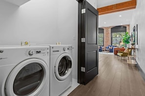 A laundry area with two front-loading washing machines, a black door, and a seating area with a sofa and chairs in the background. - Downtown NOLA Luxury Suite Hotel Rooftop Pool Sleeps 6 (New Orleans)