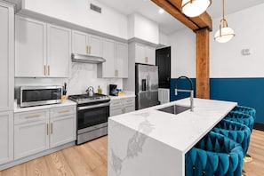A modern kitchen with grey cabinets, stainless steel appliances, a marble countertop island, and blue upholstered bar stools. - Spa Suite Bath Rooftop Pool BBQ Gym Hotel (New Orleans)