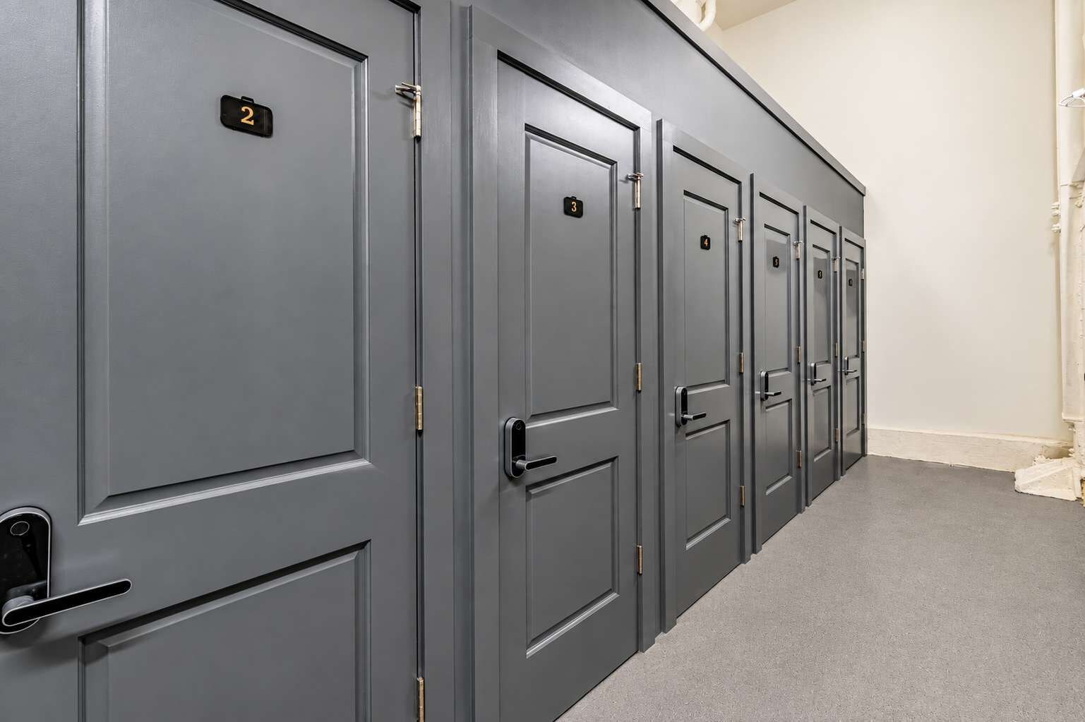 A row of grey doors with numbers indicating room assignments, a carpeted floor, and a white wall.