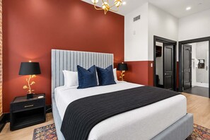Una habitación de hotel moderna con una cama grande, dos mesitas de noche, un candelabro y un baño visible a través de una puerta abierta.