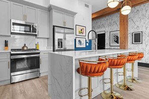 A modern kitchen with stainless steel appliances, a marble countertop, and a bar area with orange bar stools. - Lux Rooftop Pool  | Gym | Gameroom Downtown Hotel (New Orleans)