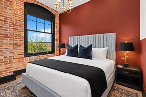 Ein Hotelzimmer mit Ziegelwand, einem großen Fenster, einem Bett mit grauem Kopfteil, zwei Nachttischen mit Lampen und einem gemusterten Teppich.