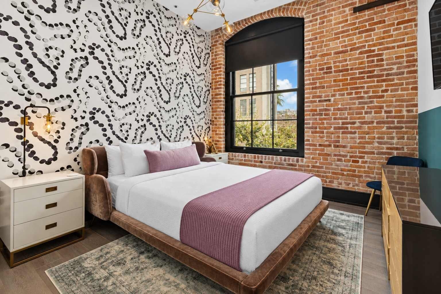 Une chambre avec un mur en briques, une grande fenêtre, un lit avec une tête de lit blanche et marron, et une table de chevet avec des tiroirs.