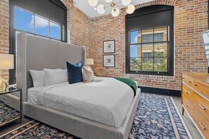 A bedroom with a large bed, a dresser, a nightstand, a window with a view, and a brick wall. - Rooftop Heated Pool Gym Gameroom Downtown Hotel (New Orleans)