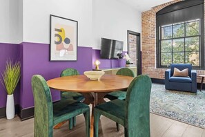 Un comedor con una mesa redonda de madera, sillas tapizadas de color verde, un cuenco blanco, una obra de arte enmarcada, un televisor de pantalla plana y una pared de ladrillo.