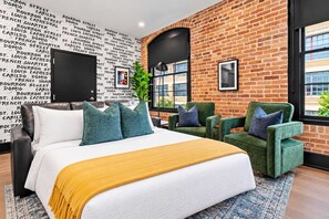 A modern hotel room with a brick wall, a large bed, green armchairs, and a black door. - Lux 2BR Suite Near Dome and FQ Hotel (New Orleans)