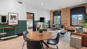 A modern hotel lobby with a round wooden table, black leather chairs, a brick accent wall, and a large window offering a city view. - Luxury 2 Bedroom Downtown Suite (New Orleans)
