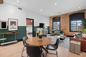 A modern hotel lobby with a round wooden table, black leather chairs, a brick accent wall, and a large window offering a city view.
