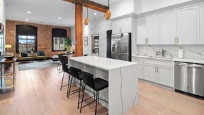 A modern kitchen with white cabinets, a central island with bar stools, a stainless steel refrigerator, and a brick wall. - Luxury 2 Bedroom Downtown Suite (New Orleans)