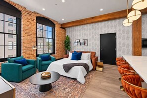 Una habitación de hotel moderna con pared de ladrillo, ventanas amplias, una cama con ropa de cama azul y blanca, sillas color turquesa y naranja, una mesita redonda pequeña y una alfombra estampada.