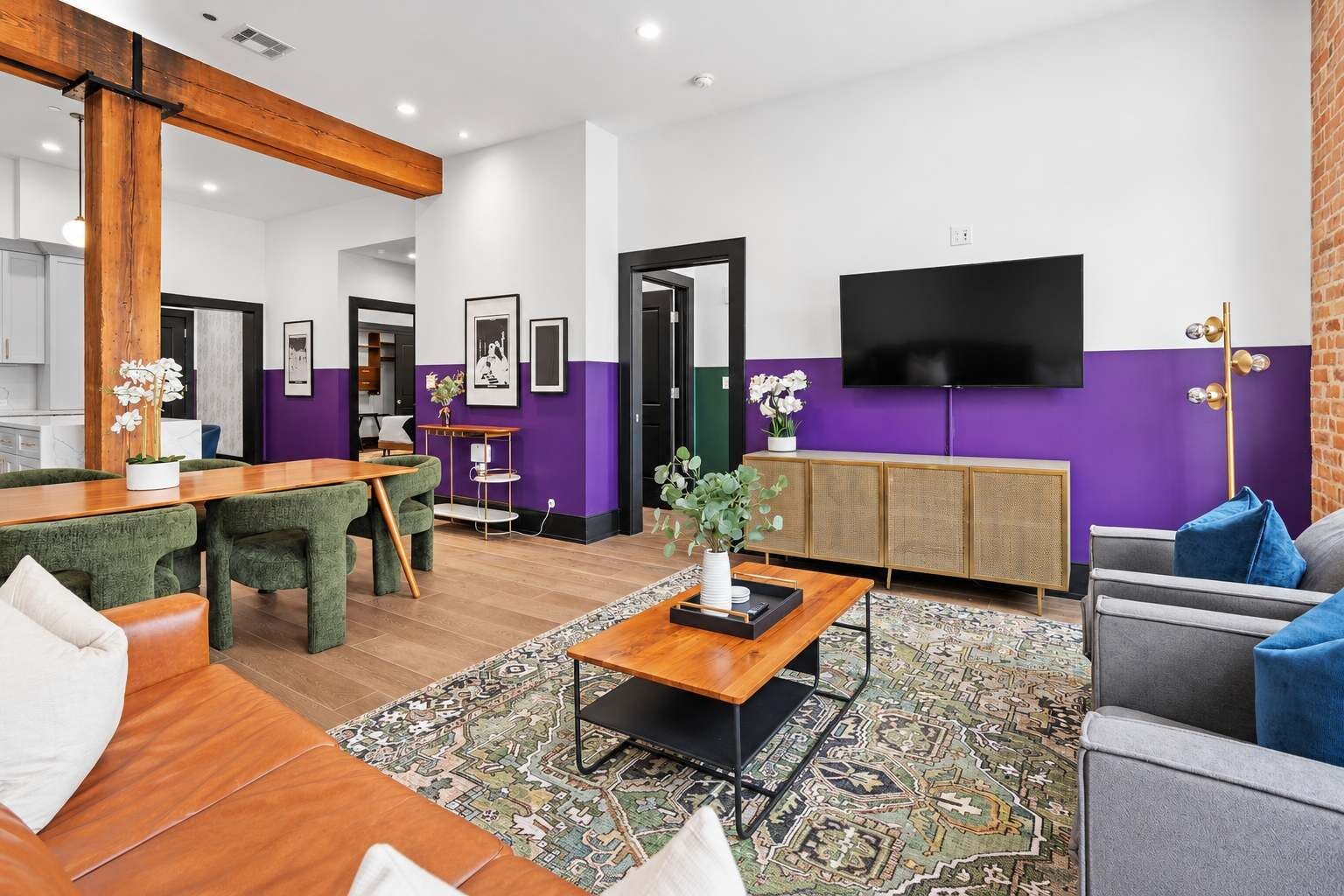 A modern living room with a wooden dining table, green upholstered chairs, a patterned rug, a grey sofa, and a flat-screen TV mounted on the wall.