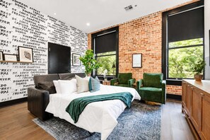 A modern bedroom with a brick wall, a large bed, a green armchair, and a view of trees through large windows. - Downtown 4BD 4BA Suite Sleeps 12 Hotel (New Orleans)