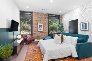 A modern bedroom with a brick wall, large windows, a teal sofa, a bed with white bedding, and a flat-screen TV. - Downtown 4BD 4BA Suite Sleeps 12 Hotel (New Orleans)