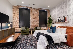 Camera d'albergo moderna con parete in mattoni, un letto grande, poltrone verdi, un tavolino e una TV.
