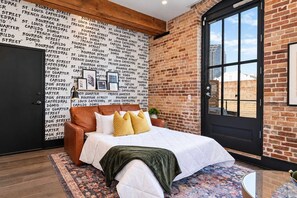 A bedroom with a brick wall, a large window, a bed with pillows, and a patterned rug.