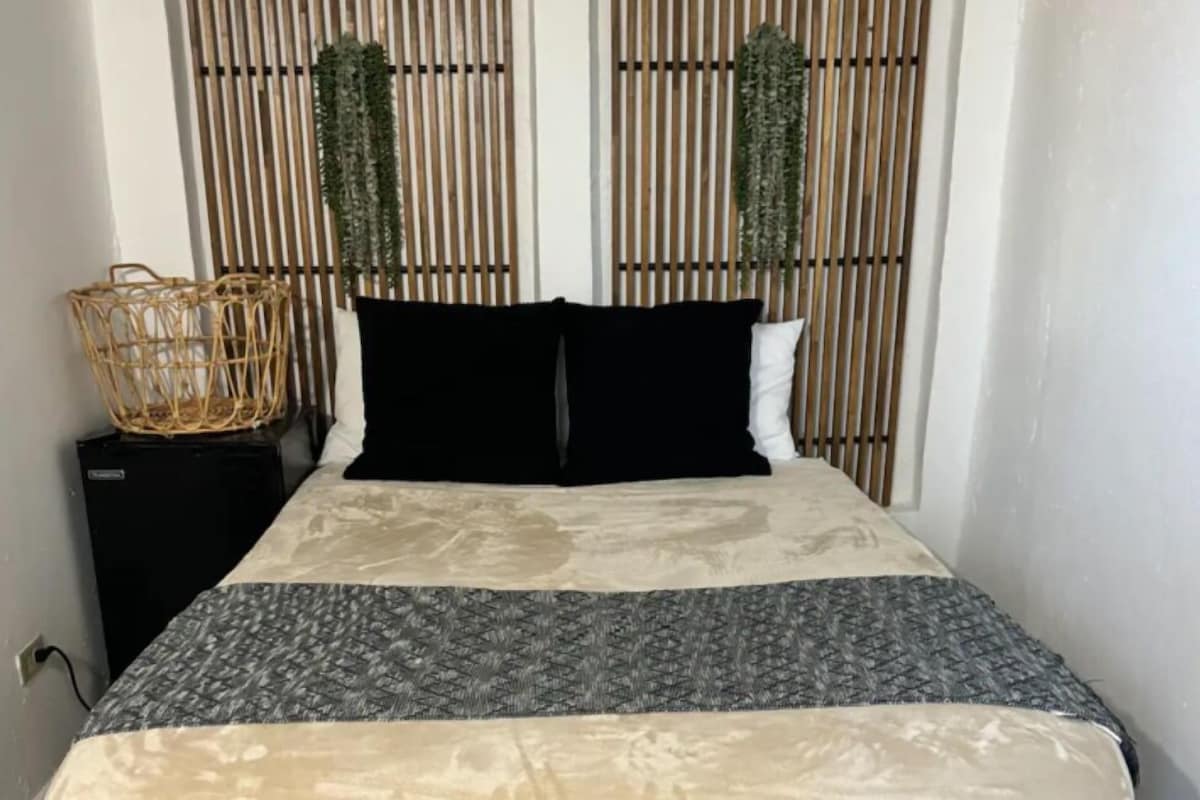 A bed with a patterned bedspread, two black pillows, and a woven basket on a nightstand.