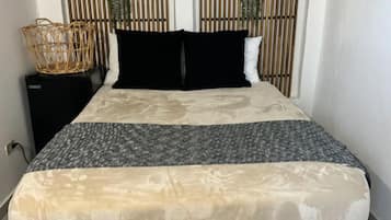 Un lit recouvert d’une couvre-lit à motifs, deux oreillers noirs et un panier en osier posé sur une table de chevet.