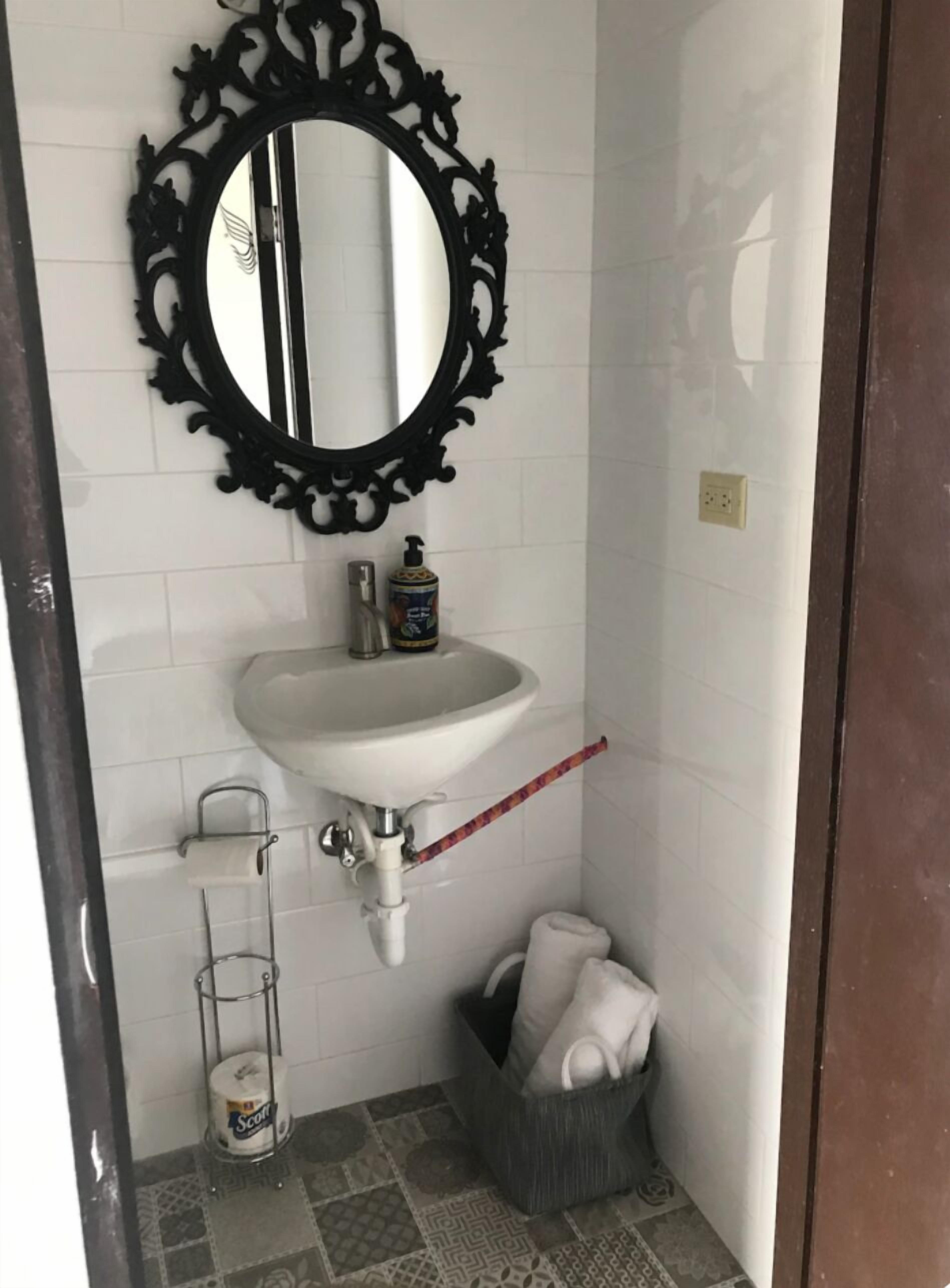 Une salle de bain avec un lavabo blanc, un miroir noir à motifs, un flacon de savon liquide, un porte-serviettes, un panier contenant une serviette et un porte-rouleau de papier toilette.