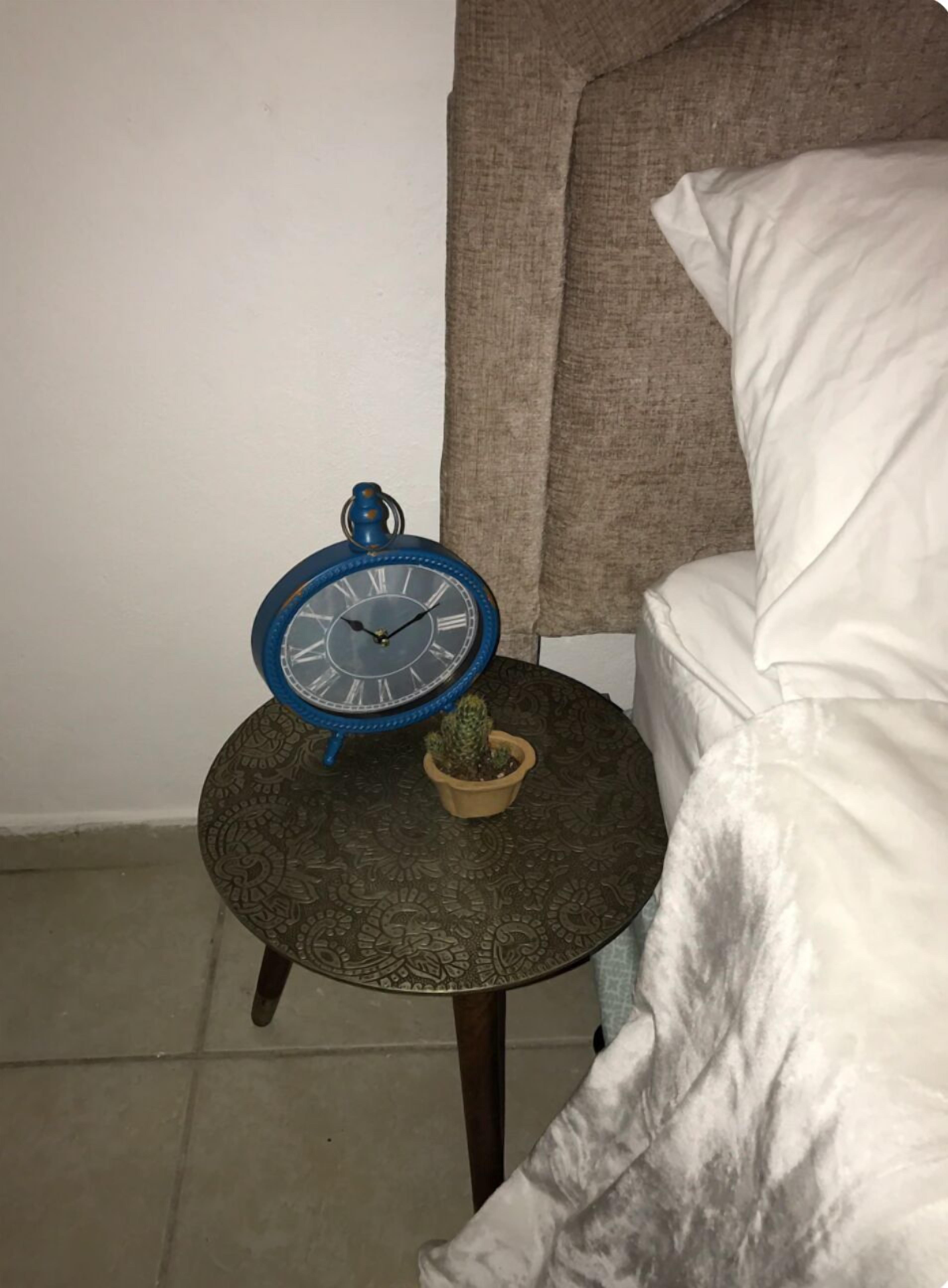 A small round table with a clock and a potted plant, placed next to a bed with white bedding.
