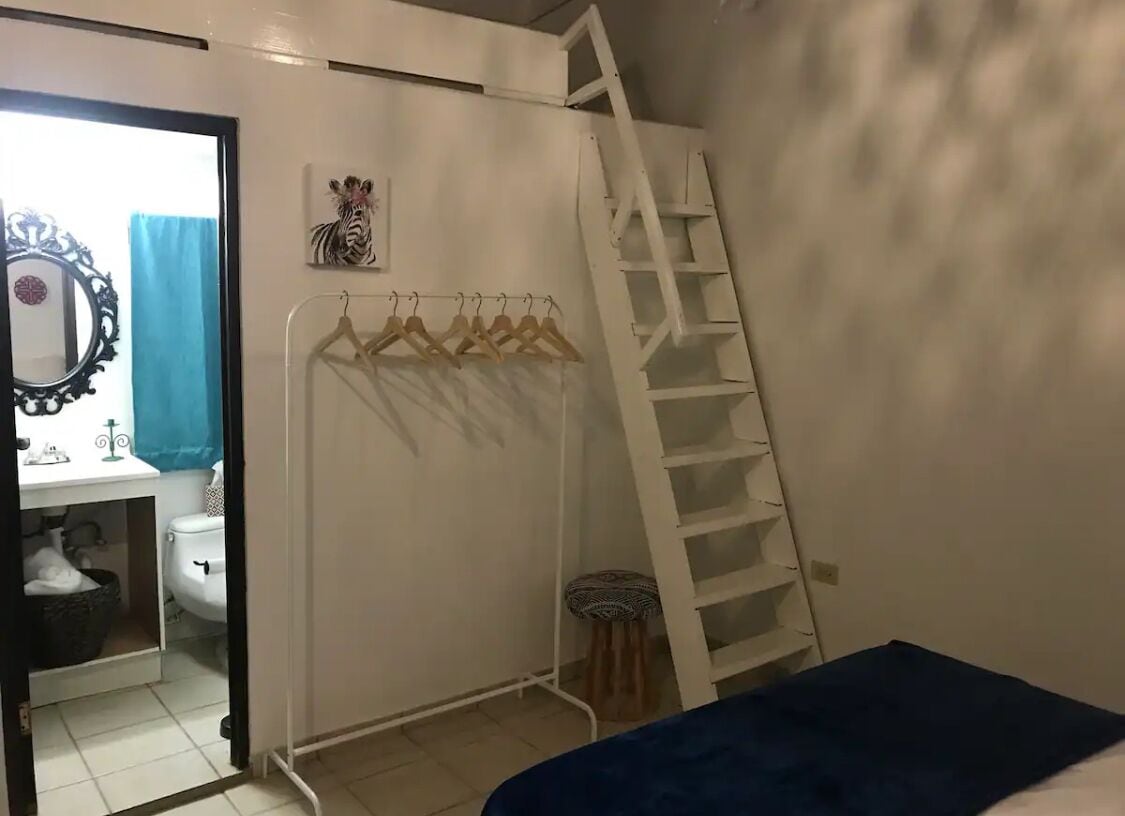 A bedroom with a bed, a wardrobe with a hanging rack, a ladder, and a bathroom visible through an open door.