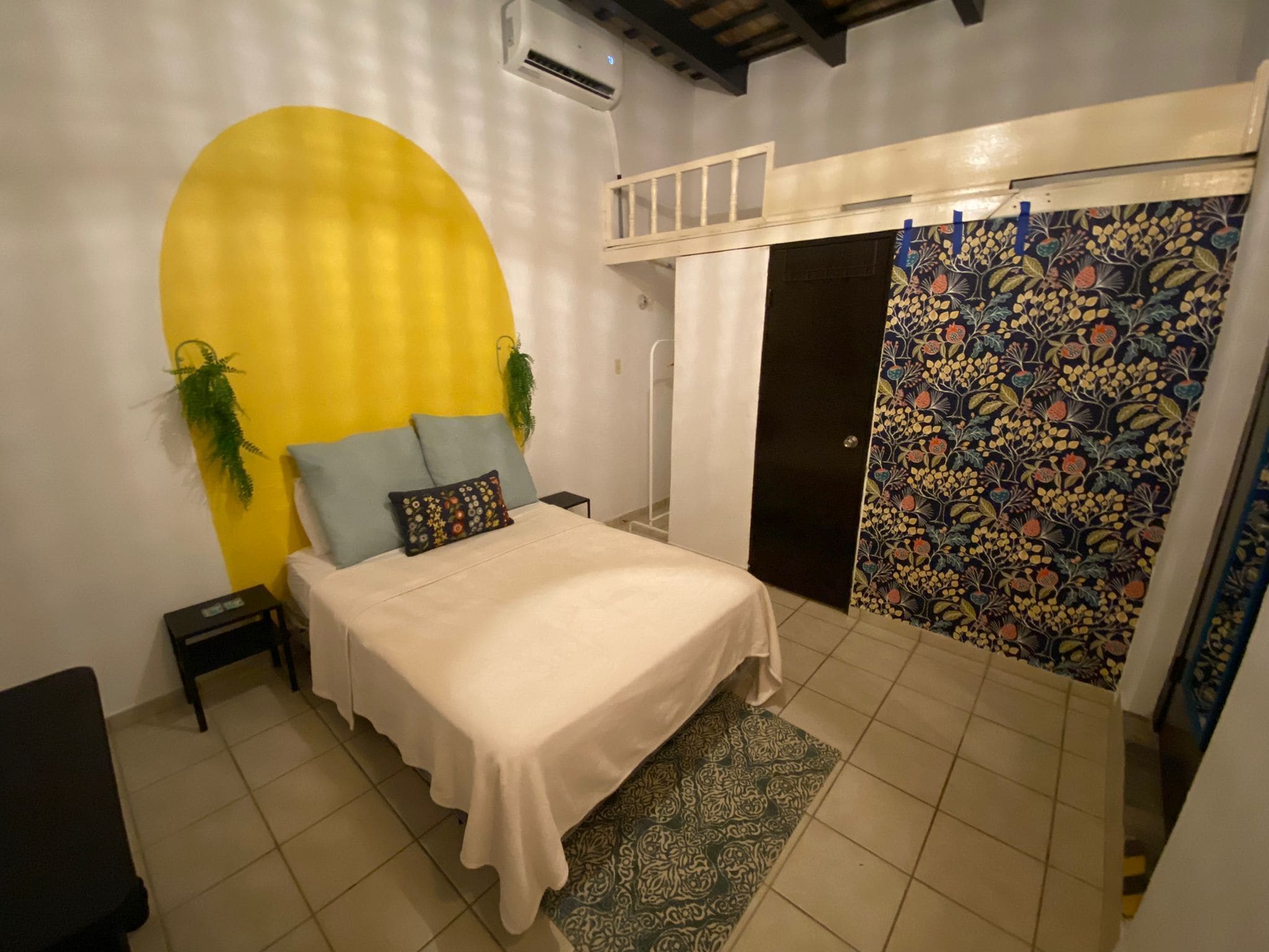 A bedroom with a bed, a nightstand, a potted plant, a wall-mounted air conditioner, and a door with a floral-patterned curtain.