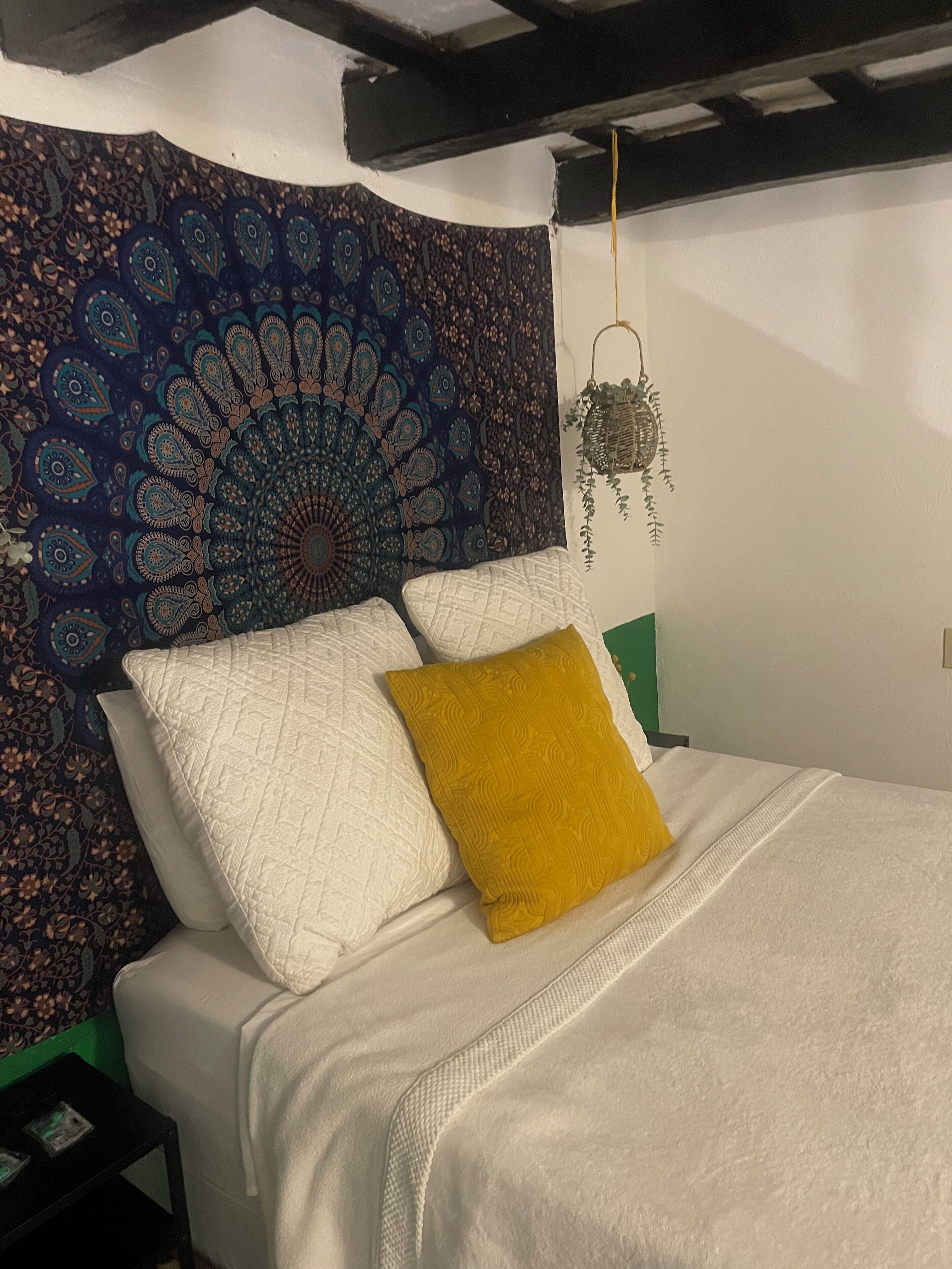 Un lit avec une literie blanche et un coussin jaune moutarde, une tapisserie décorative ornée d’un motif de paon et une lanterne suspendue au mur.