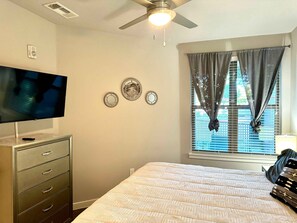A bedroom with a bed, a television on a dresser, a ceiling fan, and a window with curtains.