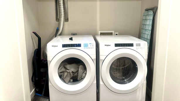 Two front-loading Whirlpool washers in a laundry room with a vent above and a vacuum cleaner to the left.