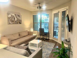 A living room with a beige sofa, a glass coffee table, a chair, a floor lamp, and a large window with blinds.