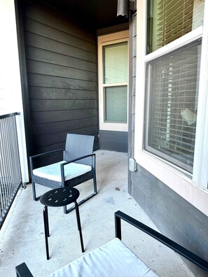 A balcony with a chair and table, a window with blinds, and a building facade.